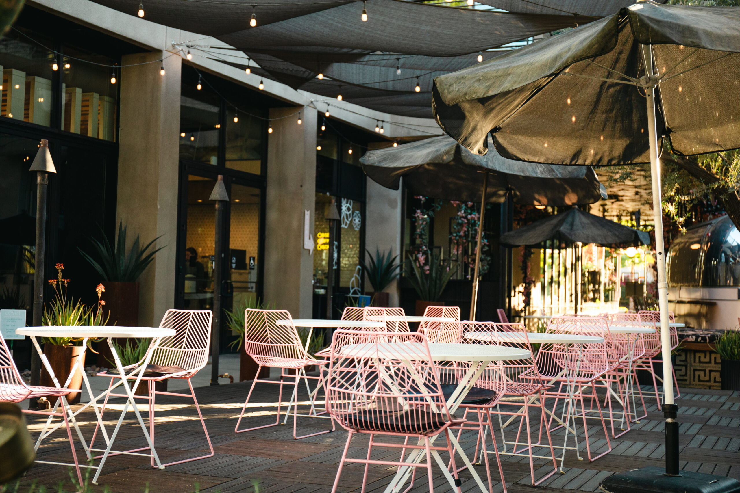 A vibrant patio exterior at Café La Jefa in Palm Springs, California. The space features ample seating with cozy tables and chairs arranged under the open sky, framed by lush greenery. In the distance, the iconic Palm Springs mountains rise, adding a serene backdrop to the inviting café setting.